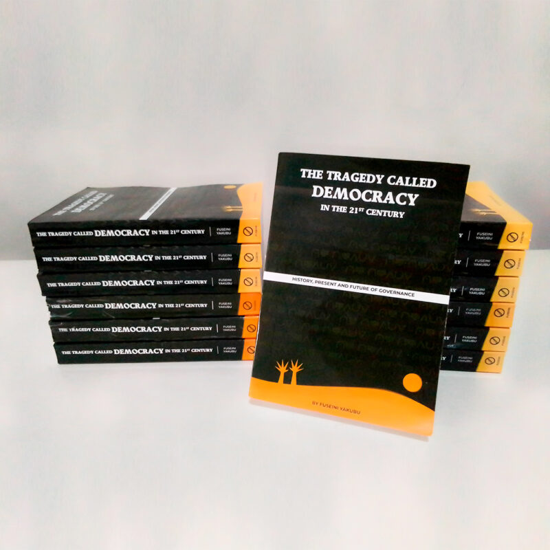12 copies of the book The Tragedy Called Democracy, stacked on top of each other as 2 stacks of 6 each, with a 1 copy leaning against the stacks